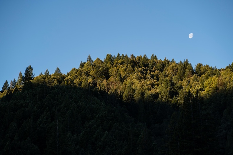 Morning sunlight crests a nearby mountain peak as the moon descends for the day.
